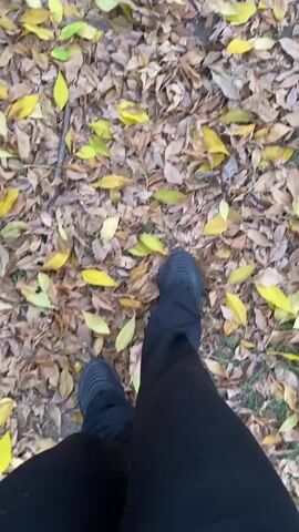 A person wearing black pants and dark shoes is walking on a ground covered in fallen autumn leaves. The leaves are mostly brown and yellow. The video is shot from a first-person perspective, showing the person's legs and feet as they move across the leaves.