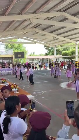 The video shows a group of children performing a dance outdoors, possibly at a school event. The children are dressed in light purple dresses and white shirts with black pants. They are dancing on a paved ground, possibly a basketball court, under a covered roof. Spectators are seated in the foreground, some of whom are recording the performance with their phones. There are decorations and tables set up to the left of the frame. In the background, more spectators are visible, as well as buildings that appear to be part of a school or community center. The event seems to be taking place during the daytime, with natural light filtering through the open sides of the covered area.