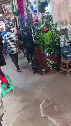 The video shows a busy marketplace or indoor bazaar. People are walking through narrow aisles lined with various merchandise displays. There is a woman crouching near a display of artificial plants and flowers. Several other individuals are walking by, including a woman in a patterned dress and a man in a black and white patterned shirt. Clothes and decorations hang overhead. The lighting suggests an indoor setting, and the environment appears to be bustling with activity.