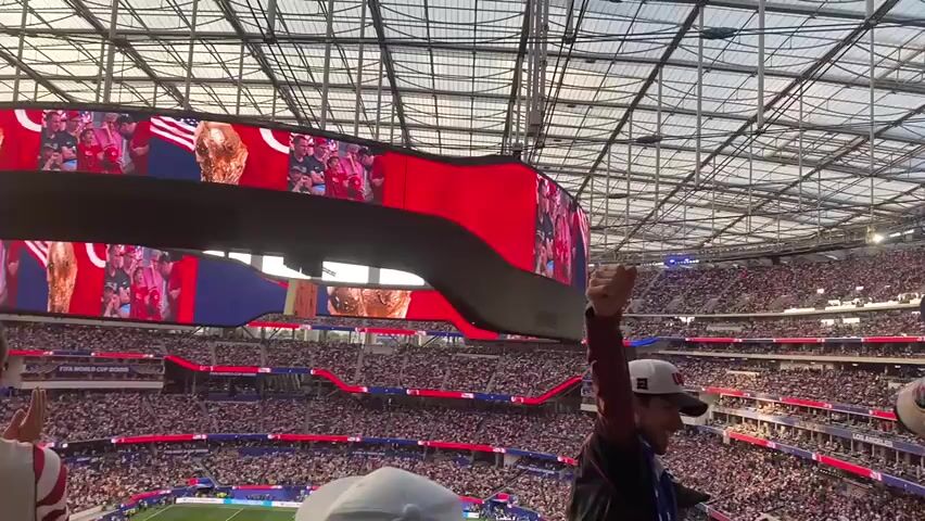 This media file captures a moment from a soccer match at SoFi Stadium in Inglewood, California. The camera is positioned in the stands, looking up at a large, circular LED screen that displays a montage of images. The screen prominently features the US flag, interspersed with photographs of people holding up a trophy, likely signifying a victory or celebration. The stadium is packed with a large crowd, creating a vibrant and energetic atmosphere. The time of day appears to be late afternoon or early evening, as indicated by the warm lighting filtering through the stadium's translucent roof. The weather seems clear. The overall mood is celebratory, with the crowd and the displayed imagery conveying a sense of national pride and excitement. Some visible text on lower screens reads "FIFA WORLD CUP 2026 LOS ANGELES".