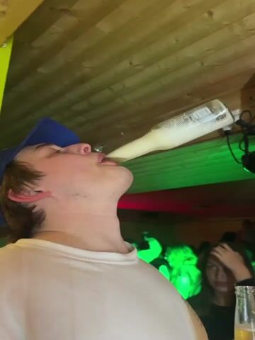 A young man, seen from a low angle, is the central subject in this video. He wears a white t-shirt and a blue baseball cap, on which white text reading "SAN ANTON" and partially "AM AM MO..." is visible. His head is tilted sharply backward as he drinks directly from a clear glass bottle containing a light yellow or off-white liquid. The bottle's white label has small, illegible black text.

The setting appears to be an indoor party or bar, characterized by a wooden plank ceiling and dim, colorful lighting. Red, blue, and green lights cast an atmospheric glow, suggesting an evening or nighttime event. In the background, other people are visible, mostly out of focus. To the right of the main subject, a woman with dark hair, wearing a dark top, holds a clear glass bottle with the word "CORONA" clearly printed on its label. Electronic equipment or lighting fixtures are mounted on the wall/ceiling in the background. The man is focused on consuming the drink.