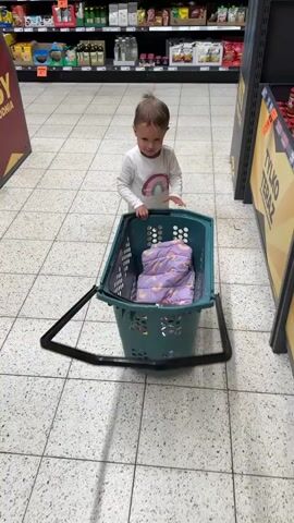 A toddler in a white shirt and pink pants pushes a green shopping basket with a handle, containing a folded patterned purple blanket. The toddler moves the basket forward in a grocery store with tiled floors, between shelves of packaged food. The sign "TYLKO TERAZ" is visible next to the shelves. The toddler smiles and appears to be enjoying pushing the basket.