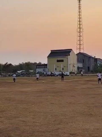 The video captures an outdoor scene at dusk, likely a sports field or an open area. In the foreground, the ground is mostly dry and brown, with some patchy grass. Several people are scattered across the field, engaged in what appears to be a casual game of soccer. Two individuals are visible in the mid-ground, one in black attire and another in white, seemingly interacting with a soccer ball. Further back, more individuals in white are spread out on the field. A yellow building with a dark roof is prominent in the background, situated behind a row of trees. To the right of this building stands a tall, lattice-like transmission tower. Several other buildings, appearing to be houses, are visible beyond the yellow structure. The sky displays a soft gradient of orange and pink hues, indicative of the setting sun. The overall atmosphere is calm and suggests a community gathering or recreational activity.