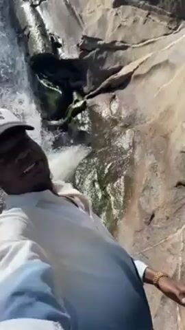 Here's a description of the video:

A man stands in front of a rocky waterfall. He is wearing a white shirt, and a white cap with a red heart that says "I LOVE HOT MOMS" on it. He is smiling and looking at the camera. Water is splashing around him. The waterfall is small and runs down a dark rocky cliff.