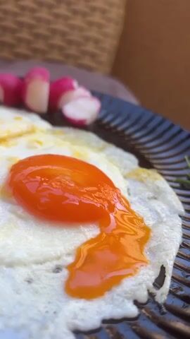 A slice of dark bread is being dipped into the runny yolk of a fried egg. The egg is on a dark plate with ridges. Several sliced radishes are in the background, along with a woven texture, possibly a placemat or basket. The egg white is cooked and white, while the yolk is bright orange and still liquid. The bread is brown and appears to be whole wheat.