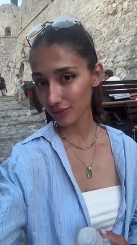 The video begins with a close-up selfie of a woman with dark hair pulled back, wearing a light blue collared shirt and a white strapless top. She has sunglasses perched on her head and is wearing two silver necklaces, one with a green pendant. She smiles at the camera, and her head is tilted slightly. The background is out of focus but appears to be an outdoor setting with stone architecture and people dining at tables. The camera movement is unsteady, indicating it's being held by the person recording. The woman blinks and smiles again, and the video continues to be a selfie with the background blurred. There are other people visible in the background, some standing and some seated at tables. The environment seems to be a lively outdoor area, possibly a restaurant or cafe.
