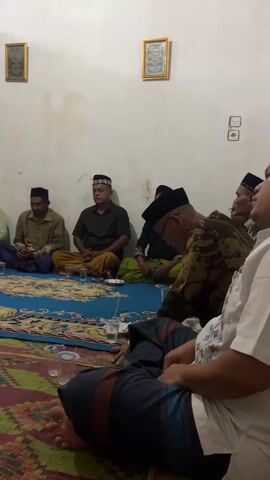 A group of men in traditional Islamic attire are seated on the floor in what appears to be a room. They are arranged in a semi-circle, facing towards the center of the room. Some men are wearing kufis or peci hats. The floor is covered with two large rugs, a blue patterned one and a green and red patterned one. There are several plastic cups scattered on the rugs. In the background, there is a white wall with two framed pictures and a doorway with a wooden door. A fan is visible near the doorway. The lighting suggests it is evening or indoors. The men appear to be engaged in conversation or listening to someone speak.