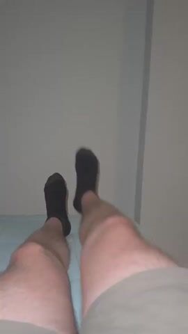 The video shows a close-up view of a person's legs and feet, which are covered in black socks with two brown stripes near the cuff. The person appears to be lying down on a light blue quilted surface. The legs are hairy, and the lighting suggests an indoor environment with a plain, light-colored wall in the background. The camera angle is from above, looking down at the legs. The video consists of several short clips, each showing a slight movement or change in perspective of the legs and feet. In some clips, the feet are crossed, while in others, one leg is extended slightly forward. The focus is primarily on the lower legs and feet.