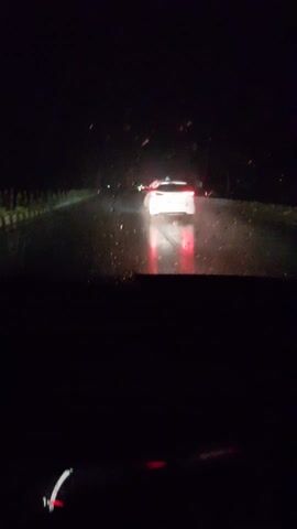 This media file captures a nighttime drive on a wet road in Paramankeni, India. The scene is viewed from inside a vehicle, with the dark interior occupying the foreground. The primary subject is a white car ahead, its taillights glowing red and reflecting brightly on the slick surface of the road. The road itself appears wet and somewhat muddy, with visible puddles and streaks of water. The surrounding environment is dark, suggesting a rural or less urbanized setting, with some faint lights and indistinct foliage visible on the sides of the road. A triangular road sign is briefly visible on the right side of the frame. The overall atmosphere is one of traveling through the dark and wet conditions, with the red taillights of the car ahead providing a focal point and a sense of movement. There is no visible text in the media file.