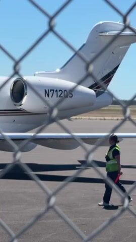 A person wearing a high-visibility vest walks on an airport tarmac beside a white private jet. The jet has the registration number N712QS visible on its tail. The person is holding a red signaling wand. The scene is viewed through a chain-link fence. The sky is clear and blue.