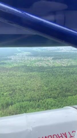 The video captures an aerial view from a small aircraft, looking down upon a green, forested landscape in Кусье-Александровский, Russia. A vast expanse of lush, green trees, appearing to be a mix of deciduous and coniferous varieties, dominates the foreground and mid-ground.

Within this verdant expanse, or bordering it, a scattered settlement comprising numerous houses with light-colored roofs is visible. These buildings are interspersed with green plots, characteristic of rural villages or small towns. Further in the distance, beyond the immediate settlement, larger clusters of buildings indicate more urbanized areas, partially obscured by atmospheric haze.

The view is framed by elements of the aircraft. A dark blue structural component, possibly a wing strut or support, runs across the top of the frame. In the lower right corner, a white section of the aircraft's fuselage or engine cowling is visible. Partially obscured text in Cyrillic script is printed on this white surface; the visible portion reads "Т АНОВ," which is likely part of a word such as "ОСТАНОВ" (STOP).

The sky appears overcast and hazy, contributing to a muted, diffused light across the landscape. No people are visible in the shot.