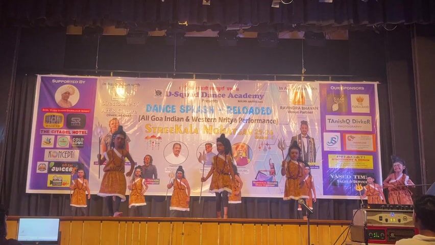 A stage performance is underway, featuring nine children dressed in Shiva costumes. The kids are evenly spaced and dance with energetic, synchronized movements. They hold trident props and some have face paint, including red markings on their foreheads.

Behind them, a large banner promotes "D-Squared Dance Academy presents Dance Splash Reloaded" with the tagline "All Goa Indian and Western Nritya Performance" and the event "Streekala Mohotsav 25-26." The banner includes logos of sponsors such as The Citadel Hotel, Koncept Juice, and several other local businesses.  

The stage is lit with spotlights. A mixing console is visible on the far right. A laptop screen is visible on the left with an individual apparently monitoring the performance. The children are the central focus.