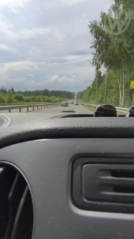 The video shows a first-person view from inside a car driving on a highway. The dashboard is visible in the foreground, with a circular vent on the left and a rectangular panel in the center. The road ahead has multiple lanes, with other cars driving in the same direction. A metal guardrail separates the lanes from the left side of the road, where there are trees and fields. On the right side of the road, there are more trees and a lamppost. The sky is cloudy.