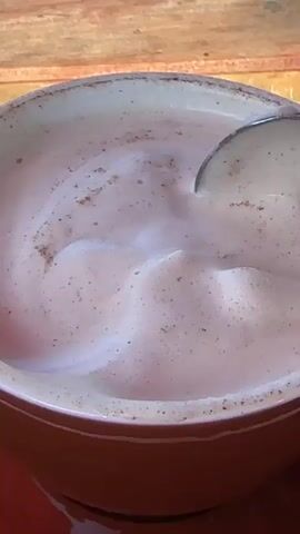 A close-up video shows a spoon stirring a frothy, pinkish-brown beverage in a brown mug with a white rim. The mug is sitting on a dark red saucer. The spoon, made of a silver-toned metal, is lifting a dollop of foam from the surface of the drink. The background is blurred and appears to be a wooden surface with warm lighting, suggesting an outdoor or rustic setting. The action is focused on the beverage and the spoon, highlighting the texture and froth of the drink. The video appears to be a short, looped clip.