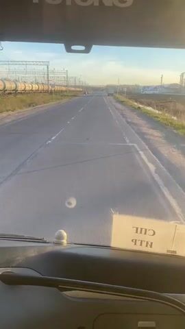 The view from inside a vehicle, likely a truck, traveling on a road. A train track runs parallel to the road on the left, with tank cars visible on the tracks. The road surface is asphalt with white lane markings. A large truck is parked on the right side of the road. The sky is blue with some light clouds, indicating it may be early morning or late afternoon. There is a small white object on the dashboard near the windshield. A piece of paper is taped to the dashboard with some text written on it in Cyrillic.