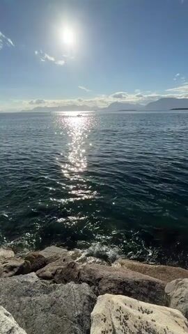 The video shows a body of water with a rocky shore in the foreground. The sun is shining brightly in the sky, creating a reflection on the water. There are mountains in the distance. A small boat is visible on the water. The waves are gently lapping against the shore. The water is dark blue and clear. The sky is a bright blue with a few scattered clouds. The rocks are varied in color and texture.