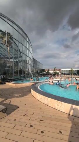 The video shows a crowded outdoor swimming pool area with a large glass-enclosed building on the left. The pool is circular with people swimming and relaxing in the water. A wooden deck surrounds the pool. The sky is cloudy. There is a shadow of a person holding a phone in the foreground. The overall scene depicts a resort or water park on a partly cloudy day.