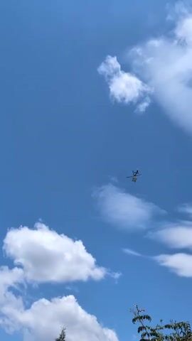 A helicopter flies across a clear blue sky dotted with white clouds. The helicopter is small in the frame, making it difficult to discern specific details. The camera angle is looking up at the sky from a ground perspective, with the top of some trees visible at the bottom of the frame.