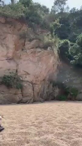 Here's a detailed description of the video:

The video shows a beach scene on a sunny day. It opens with a wide shot of a rocky cliff covered in vegetation, with two people lying on towels in the foreground, sunbathing on the sand. The camera then pans up and away to reveal a wider view of the beach, coastline, and the ocean. A couple of birds are visible walking on the sand near the shoreline. The camera eventually focuses on the perspective of someone lying on the beach, capturing the view of the water and coastline between their legs. There are boats in the distance on the water. The coastline is green with vegetation and includes some houses. The sky is blue with some clouds.