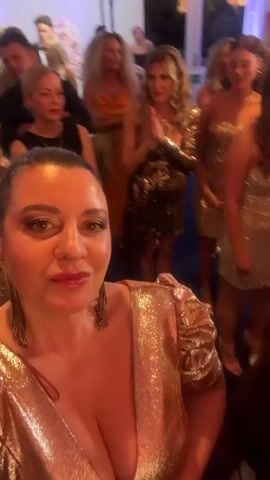 The video shows a woman in a sparkly gold dress, taking a selfie at what appears to be a party or event. She has dark hair pulled back in a ponytail, and is wearing dangling earrings.  Behind her are numerous other women, many of whom are also wearing sparkly gold dresses. The lighting is bright, and there appears to be photography equipment set up. The setting appears to be a banquet hall or similar venue.