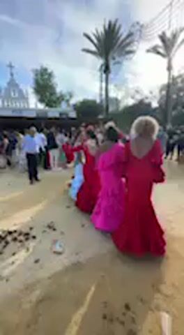 This video captures a lively outdoor scene at what appears to be a festival or fairground in Jerez de la Frontera, Spain. The setting is bright and sunny, with a blue sky and some white clouds, suggesting it is daytime. The ground is sandy or dusty.

The main subjects are several women dressed in vibrant, traditional Spanish attire, characteristic of flamenco or regional festivals. They wear long, flowing dresses with ruffled skirts in striking colors, primarily shades of red, fuchsia pink, and light blue. The dresses feature full sleeves and V-necklines.

From 0:00 to 0:02, the video shows several women from behind, moving or dancing, with palm trees and distant white buildings in the background. From 0:02 to 0:05, the camera briefly focuses on three women from the waist up. They are adorned with traditional accessories including mantones (embroidered shawls) draped over their shoulders and flowers in their hair, adding to the festive look. Their expressions are calm, appearing to be observing or posing.

From 0:05 to 0:11, the camera captures a wider view of the group of women, who are dancing or posing together. Overhead, festive string lights and decorations are visible, strung across the area, further indicating a celebratory event. The atmosphere appears joyful and festive, with many other people subtly visible in the background. No visible text is present in the video.