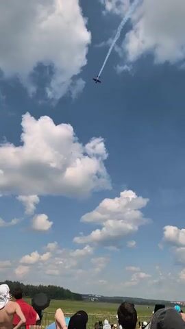 The video shows a crowd of people gathered in a grassy field, watching an airplane perform aerobatics in a blue sky with white clouds. The airplane is small and dark-colored, leaving a white smoke trail as it ascends at a sharp angle. The spectators are standing and looking up at the sky, some with their hands raised. One person in the foreground is wearing a white t-shirt and blue shorts, with a white bandana on their head. Another person is wearing a black tank top and a floral skirt. There are tents set up in the background, suggesting an outdoor event or air show. The overall scene depicts a sunny day with a focus on the aerial performance.