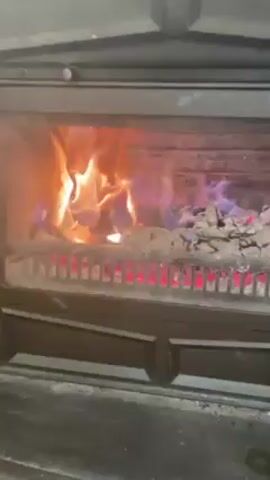 Inside a black wood-burning stove with a glass door, flames are burning in the firebox. The fire is mostly orange, but there are sections of blue flames. The logs are on a grate above a layer of hot red embers and ash. The camera remains still. There is some indistinct speech audible in the background.