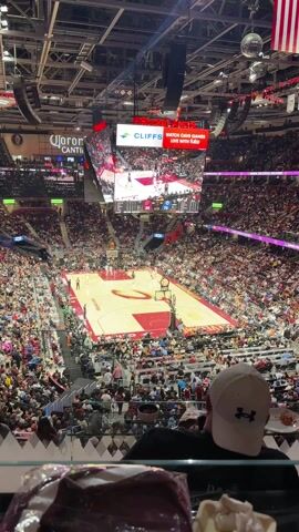 The video shows a basketball game from a high angle, potentially from the stands. The court is maroon and beige with a large "C" logo in the center. Two teams in red and white jerseys are actively playing. A large digital scoreboard hangs above the court, displaying the score and other game information. The arena is filled with spectators in various clothing. The viewer's perspective is partially obstructed by a person in a white baseball cap, sitting in front of the camera. A container with some food is visible near them.