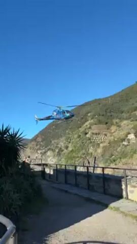 A blue and black helicopter flies against a clear blue sky and past a green hillside. A thin cable hangs down from the helicopter. A gray stone walkway with a black metal fence sits below the helicopter. There are green plants on either side of the walkway. Three people are standing near the plants.