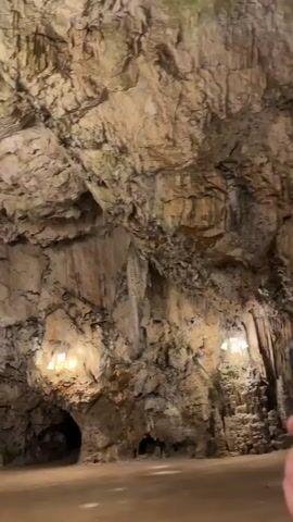 The video shows a panning shot of a large, dimly lit cave. The cave walls are made of rock formations, some of which are illuminated by artificial lights. There are also stalactites hanging from the ceiling. The floor of the cave appears to be made of dirt or sand. No people are visible in the video.