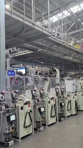 The video shows an indoor factory setting, likely a manufacturing facility. The foreground is dominated by a line of automated machines, primarily white with some black accents and digital displays. These machines appear to be part of a production line, with hoses and cables connected to them. Above these machines, a complex network of robotic arms can be seen, some of which are actively moving and interacting with components within a protected enclosure. The overall impression is one of advanced automation and industrial production.

The scene is brightly lit, suggesting it is daytime or the facility has extensive artificial lighting. The architecture of the building is visible in the background, with a high ceiling, exposed metal structures, and industrial-looking ventilation systems. There are no visible people in the immediate foreground, though there might be some activity further back or within the robotic enclosures, obscured by the machinery. The mood is sterile and functional, typical of an industrial environment focused on efficiency and automation.

Visible text includes a sign that reads "MB-10" above one of the robotic enclosures. Some of the machines in the foreground also have labels and digital readouts, though the text is too small to be legible. The overall context of a manufacturing plant in Yufengshan, China, suggests this could be a facility producing consumer goods, electronics, or automotive parts, given the nature of the machinery.