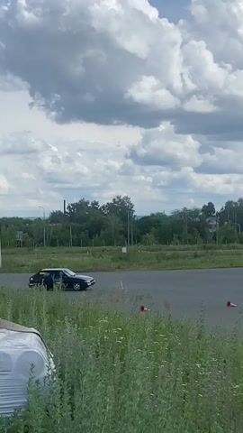 The video shows a dark-colored car driving on a paved track. The track appears to be a race track or a similar venue, with orange and white cones placed around the course. In the background, there are trees, buildings, and a cloudy sky. The car is the primary focus of the video, and it seems to be performing some kind of maneuver, possibly a drift or a turn. There are no visible people inside or immediately around the car. The overall environment suggests an outdoor event or practice session related to motorsports.