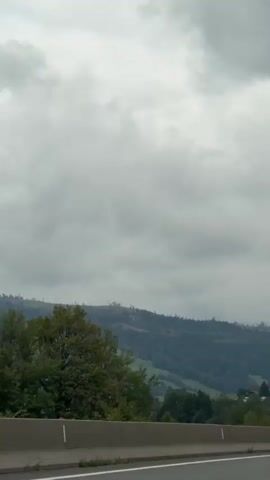 The video captures a highway view with a cloudy sky and a mountainous landscape in the background. The highway has a concrete barrier on the right side and white lane markings. Several cars and a truck are visible driving on the highway. The landscape consists of green hills and trees. The weather appears overcast.