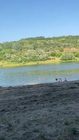 This video depicts a leisurely day at a sandy riverbank in Dzerzhinskoye, Moldova. The sun is shining brightly, indicating it's daytime, likely mid-morning or early afternoon, with a clear blue sky overhead. The river is calm and reflects the light, bordered by a lush, green, tree-covered hillside.

Several people are enjoying the water and the beach. In the water, some individuals are swimming or playing, with one person appearing to be wearing a flotation device. Closer to the shore, a small group of people are sitting on the sand, some seemingly relaxing or conversing.

On the beach, there's a wooden umbrella providing shade, under which a person is seated. Nearby, a small blue kayak is pulled up onto the sand, along with a pink bag, suggesting preparation for or rest from water activities. The sandy foreground of the beach appears somewhat disturbed, with footprints and possibly tracks from small creatures.

The overall atmosphere is one of relaxation and enjoyment of a warm, sunny day in a natural setting. The location cues like the sandy bank, river, and the vegetation on the hillside are consistent with a riverside recreation area in a place like Dzerzhinskoye. There is no visible text in the video.
