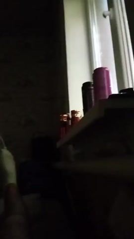 A fan is on a windowsill and is blowing air. There are several cans and a bottle on the windowsill. The room is dark.