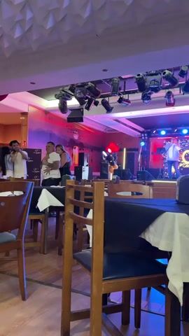 The video shows an indoor setting that appears to be a restaurant or a similar establishment. There are tables with white tablecloths and chairs, suggesting a dining area. In the background, there is a stage with a band performing. Several people are visible, some standing and interacting, while others are on the stage. The lighting is vibrant, with colored spotlights illuminating the stage and the performers. The overall atmosphere seems lively and social.
