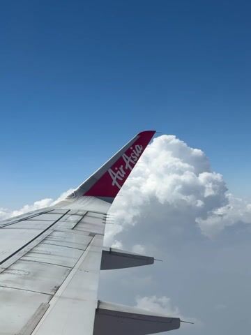 From an airplane window, the wing of an AirAsia plane is visible against a backdrop of fluffy white clouds and a clear blue sky. The AirAsia logo is prominently displayed on the wing tip. The plane appears to be in flight.