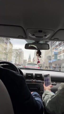 The camera is positioned inside a car, looking out through the windshield towards a city street in Europe. A person in the driver's seat is seen from the shoulder down, focusing on driving. To the right, a passenger holds a tablet displaying a map. The car is stopped at a red light behind a white SUV. Buildings line the street, including one with a "BOSCH SERVICE" sign. In the distance, the spires of a church are visible. A small air freshener dangles from the rearview mirror. Pedestrians are seen walking on the sidewalks. The weather appears overcast.