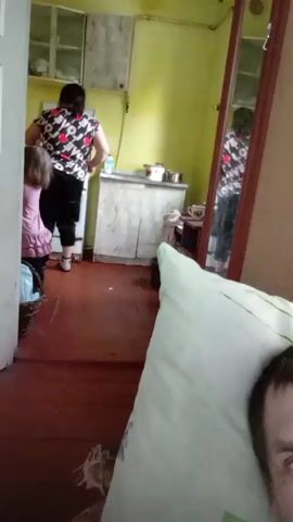 The camera is situated on a bed, likely in a bedroom, as evidenced by a pillow in the foreground. The view looks into an adjacent room, possibly a kitchen. A woman in a patterned top and black pants stands near a counter in the kitchen, appearing to be cooking. A young girl in a pink dress stands beside the woman. The walls of the kitchen are painted a light green color. The floor is reddish-brown and appears to be made of wood. A mirror on a wooden frame reflects the woman in the kitchen.