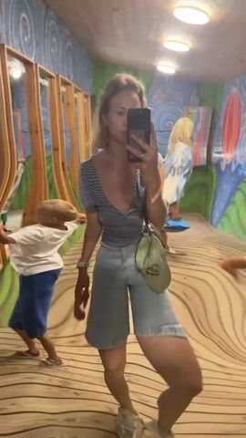 A woman with blonde hair tied back in a ponytail is taking a selfie in a room with a distorted mirror. She is wearing a striped t-shirt, denim shorts, and white sneakers. She is holding a smartphone in her right hand and a brown bag in her left hand. A young boy with blonde hair is visible in the mirror behind her, also distorted. The room has wooden floors and walls with colorful murals. The mirror reflects the scene in a funhouse-like manner, elongating and compressing the figures and the surroundings. The lighting in the room appears to be from overhead fixtures.