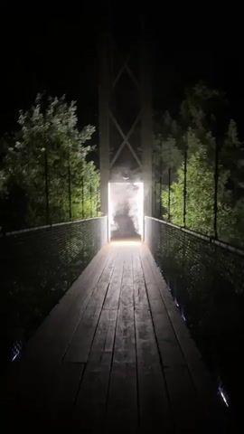 A nighttime video shows a wooden pedestrian suspension bridge. The bridge has a central tower with an illuminated opening at the top. Smoke or fog is visible in the opening and on the bridge. Trees line the sides of the bridge. The video appears to be a still image with a slight flicker effect.