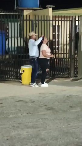 A man and a woman are dancing to music in front of a gated house at night. The man wears a baseball cap, a light blue button-down shirt, blue jeans, and white sneakers, and is facing the woman, holding a beer bottle and occasionally touching her head. The woman is wearing a light peach-colored shirt, black pants, and white sneakers. Next to them is a yellow cooler with a black handle. Behind the fence, there is a house with a blue water tank on the roof. To the side, there is a group of people sitting under a shelter, out of focus in the background. The street appears to be made of asphalt.