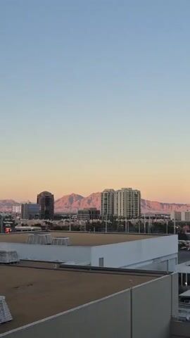 The video shows a panoramic view of a city landscape during sunset. In the background, there are mountains bathed in the warm hues of the setting sun. Several tall buildings dot the cityscape. In the foreground, there's a large flat roof, appearing to be part of a building, with some white objects stacked on it. As the camera pans from right to left, a massive, spherical, red structure, resembling a dome, comes into view. This structure has bright white text on it, which is partially visible. The sky transitions from a light blue at the top to a pale orange closer to the horizon.