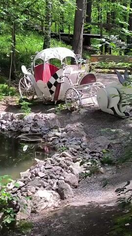 The video shows a decorative, white, horse-drawn carriage with red upholstery and a checkered pattern on its side, positioned in a wooded area near a pond. The carriage is stationary. To the right of the carriage, a large, white, abstract sculpture resembling an elephant's rear is partially visible. The pond is surrounded by rocks and vegetation. Sunlight filters through the trees, creating dappled shadows. The overall scene appears to be an outdoor decorative setting, possibly a park or garden. There are no people or animals actively interacting with the objects in the scene.