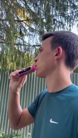 This is a short video clip showing a young man in an outdoor setting. He is wearing a teal t-shirt with a white Nike logo. The man is holding a pink electronic device with a digital display to his mouth and appears to be exhaling a plume of white vapor. He then looks up towards the sky and back towards the camera with a slight smile.

The scene appears to be daytime, with sunlight filtering through the branches of trees overhead, suggesting it is likely a pleasant day. Behind the man, there is a corrugated metal fence and further in the background, hints of trees with yellow and orange foliage, possibly indicating an autumn season. The overall setting suggests a casual, outdoor environment, perhaps a backyard or park. The man's expression is relaxed and he seems to be enjoying the moment. There is no visible text in the video.
