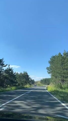 This is a first-person perspective video of driving on a paved road through a natural landscape. The road is asphalt with double white lines marking the lanes. Tall, green pine trees line both sides of the road, creating a shaded canopy in some sections. The ground on the sides of the road is covered in green grass and some rocky terrain. In the distance, there are rolling hills and more trees. The sky is a clear, bright blue with a few wispy white clouds. The sun appears to be high in the sky, suggesting it is daytime, likely midday.

There are several vehicles visible on the road. Closer to the camera, a blue car is parked on the left side of the road, and two people are standing next to it. Further down the road, other vehicles can be seen traveling in the same direction as the camera. One appears to be a small white car or motorcycle. The overall mood is peaceful and scenic, suggesting a leisurely drive through a rural or mountainous area. The location context provided is Cajetina Municipality, Serbia, which aligns with the mountainous, forested scenery. No visible text is present in the video.