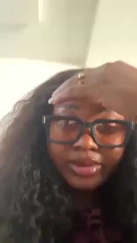 A woman with dark curly hair and glasses rests her hand on her forehead. She is wearing a pink top and a ring on her finger. The background is a plain white ceiling. The woman looks directly at the camera.