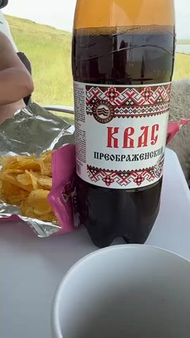 The video shows a close-up of a table with food and drinks, likely outdoors given the background scenery. A large plastic bottle of dark liquid, possibly kvass, with a label featuring red and white ethnic patterns, stands prominently. To its left, a bag of potato chips is opened, revealing a pile of golden chips. Another plastic bottle, this one containing a pale yellow, bubbly liquid, likely beer, is visible. The camera pans slightly, capturing the details of the drinks and chips. The overall scene suggests a casual outdoor gathering or picnic.