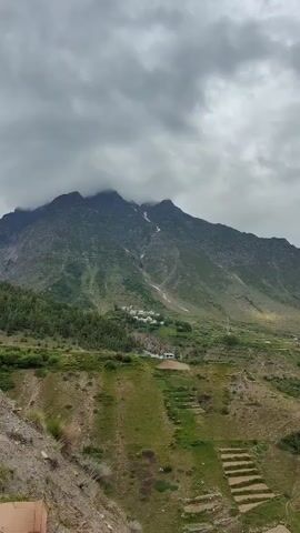 A panoramic shot pans across a valley with steep, rocky mountainsides. The sky is overcast with thick clouds. In the mid-ground, terraced fields are visible on the slopes, some appearing cultivated with greenery. A small village with scattered buildings is nestled further up the valley. A river or stream snakes through the valley floor, its color appearing murky brown. The foreground consists of a rocky, dirt embankment with sparse vegetation. The overall impression is a rugged, rural mountain landscape under a cloudy sky.