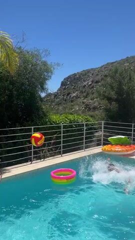 A small swimming pool is bordered by a tiled deck and a low white wall. A palm tree stands beside the wall. A black inflatable tire inner tube floats in the pool, with the words "HIGH SPEED RADIAL TIRE" printed on it. A small orange and yellow ball is near the tube. Someone throws the ball into the water, creating a splash.